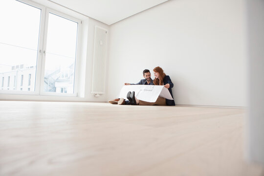 Young couple sitting on floor of their new flat, looking at ground plan
