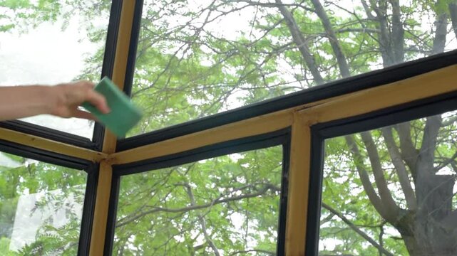 Close-up interior view of woman hand and forearm wiping dust and grime from balcony window frame with green sponge, cleaning sill, corner joints, seal edges, and painted surface in soft daylight