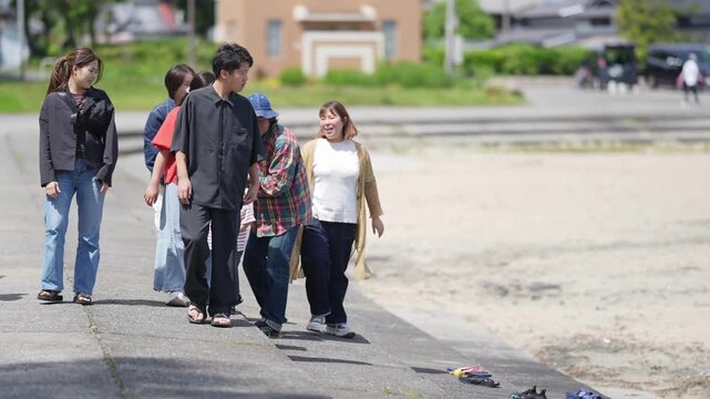 Group walks together along a paved path. Children play near adults, sharing smiles. Natural daylight illuminates the scene. Urban park setting with buildings and greenery