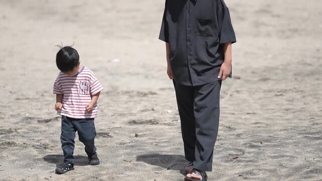 Young boy walks beside adult on sandy beach. Child looks down, curious about ground. Adult strides calmly, guiding child. Scene captures simple, peaceful beach moment. Ideal for family, nature