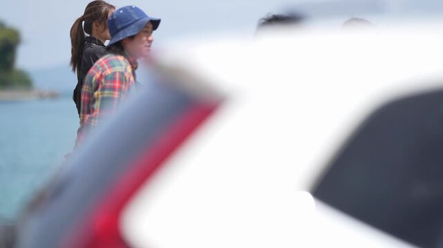 Family strolls near lake, enjoying sunny day. Young child smiles, held by parent in plaid shirt. Dog walks beside them, leash in hand. Vehicle window shows blurry reflection of group