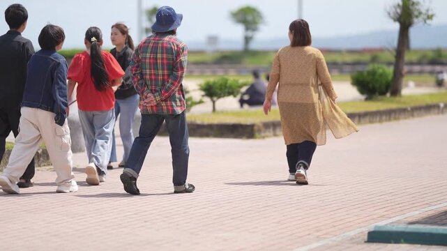 Sunlit park path, people stroll together. Group walks side by side, chatting, laughing. Woman in tan coat walks alone, calm. Perfect for travel, leisure, or urban lifestyle scenes