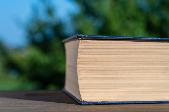 Closeup of thick closed book with yellowed pages and blue spine resting on wooden surface against blurry soft green nature and blue sky background