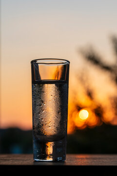 Cold vodka shot glass with water drops on sunset bokeh background, closeup. Chilled, misted shot glass filled with transparent alcohol or vodka stands on wooden surface against warm golden sunset sky