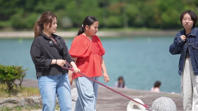 Two women walk a white dog along a lakeside path. One woman holds the leash, guiding the dog gently. Bright red top contrasts with calm lake waters behind them. They stroll with ease