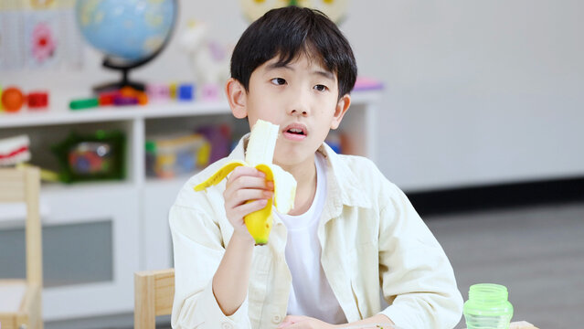 Young Asian Boy Eating a Banana During Snack Time in Classroom