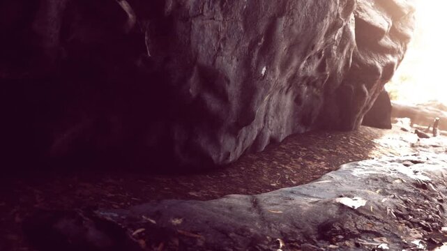 People walk through a cave with sunlight coming in from the entrance. The rocks surround the ground. Leaves are scattered on the pathway inside the cave.