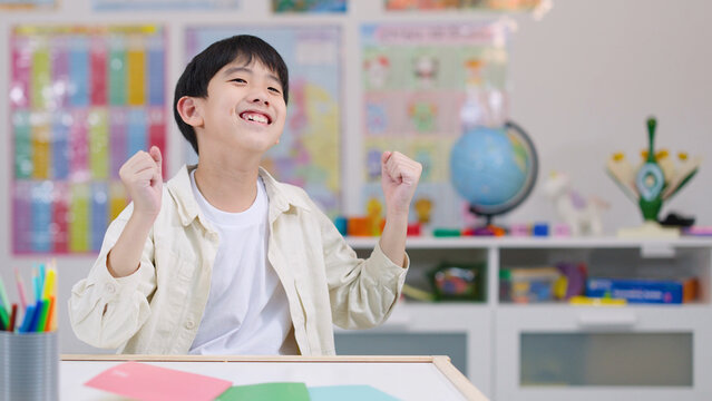 Cheerful Asian Schoolboy Raising Fists in Celebration at His Classroom Desk