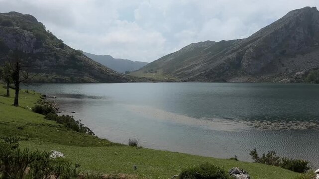 Breathtaking summer landscape of a serene mountain lake surrounded by lush green valleys, rocky peaks, and dramatic cloudy skies in Picos de Europa National Park, Spain. 
