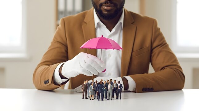 Professional man in a brown suit holding a pink umbrella over a group of tiny business people on white table