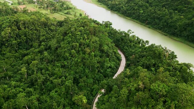 Picturesque Aerial flyover of a winding road cutting through lush tropical forest hills beside calm Pajo River - Virac, Catanduanes, Philippines