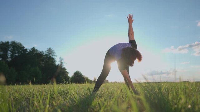 A woman stretches in a grassy field, engaged in sport. The woman moves gracefully under the bright sky, emphasizing her dedication to sport.