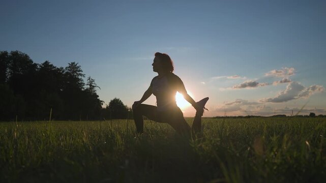 A woman stretches in a field at sunset. The woman poses to enhance flexibility while enjoying the peaceful scenery in the open air.