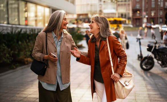 Two mature women, dressed in a tan blazer and an orange leather coat, face each other mid stride on a city sidewalk, one gesturing expressively while both smile, with a blurred street scene behind