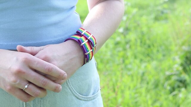 Woman in light blue top with rainbow LGBT bracelet on wrist against blurred green background. Close-up of young girl's hands with bright rainbow textile pride strap, tolerance and summer concept