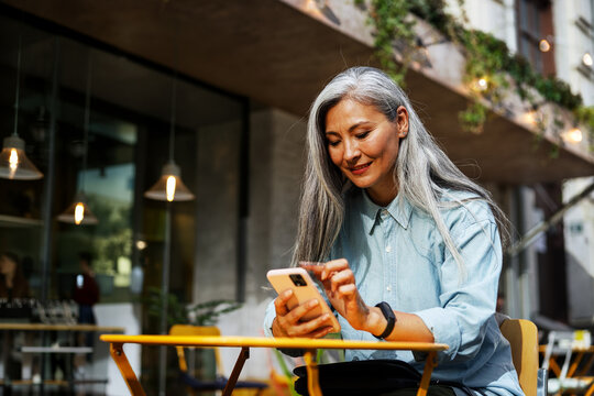 A mature woman with long gray hair, wearing a light blue denim shirt and a smartwatch, scrolls on her smartphone at a yellow cafe table outdoors, with warm pendant lights and trailing vines visible