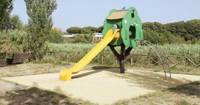 A colorful children's playground slide structure shaped like a green treehouse with a yellow slide and climbing arch, set on a padded play surface outdoors surrounded by green trees under a sunny sky.