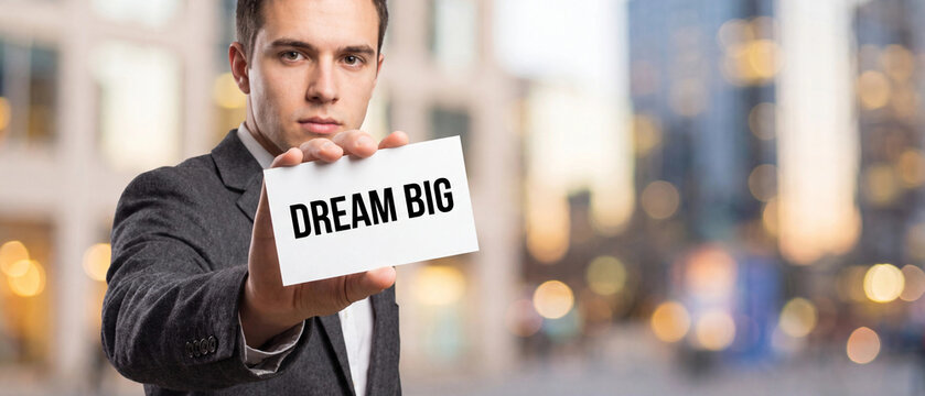 Businessman holding a sign with the text dream big in front of a blurred city background