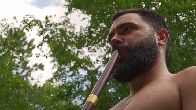 Bearded man playing a traditional wooden didgeridoo outdoor. Ethnic music performance and sound healing meditation in a summer forest.