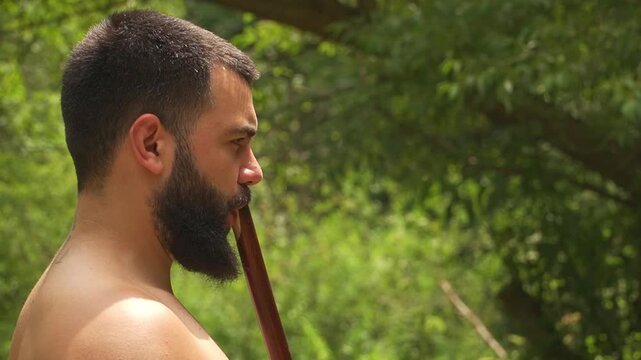 Bearded man playing a traditional kaval flute. Long pipe wooden wind instrument performance in a green forest.