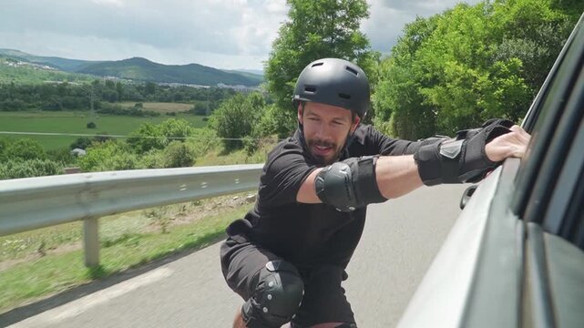 A man in protective gear rollerblades while holding onto a car, traversing a rural road with lush greenery.