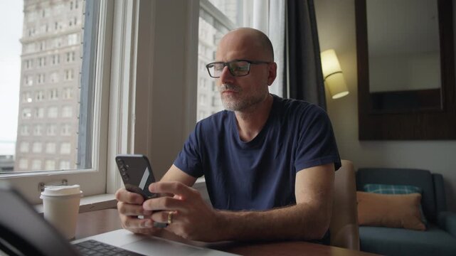 Man checks messages on smartphone, smiles, and continues working on laptop in a home office with city view.