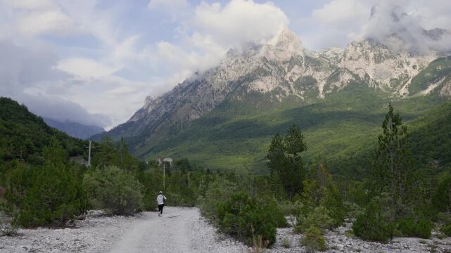 A lone hiker walks down a rocky trail surrounded by lush greenery, framed by towering mountains under a partly cloudy sky in Valbona Valley, Albania.