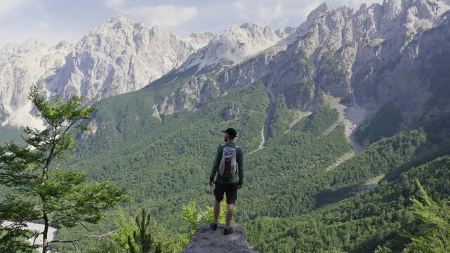 A lone hiker stands on a rocky outcrop gazing across the lush green Valbona Valley beneath towering jagged peaks in Albania under soft daylight.