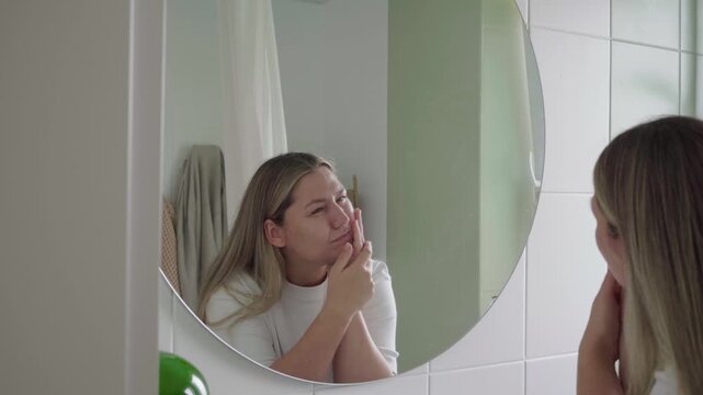 At a bathroom mirror, a long haired person opens their mouth and presses a cheek, reacting to sudden toothache.
