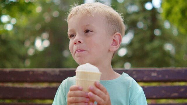 Cute little blonde boy sitting on a park bench and biting an ice cream cone. Kid eating a white ice cream on a bench in a bright summer park.