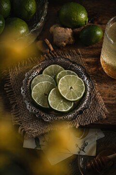 Close-up of lime slices on an ornate metal plate surrounded by burlap, fresh limes, and ginger on a wooden table with warm, moody lighting.