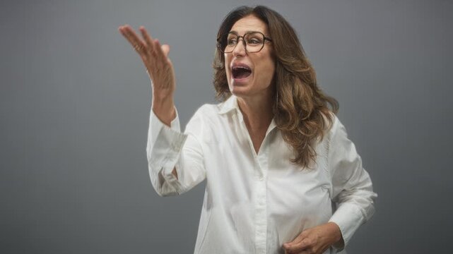 Woman gesturing with hands and open mouth while speaking in a studio, wearing glasses and a white shirt; surprise spontaneity conversation.