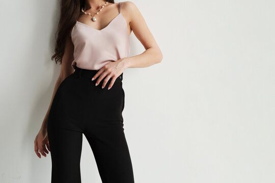 A faceless woman wearing a pink satin camisole and high-waisted black trousers on a light background with empty copyspace on the right.