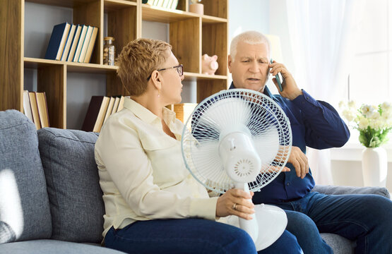 Seniors couple heatwave overheating fan phone home repair. They contact a technician about failed cooling in summer and try to cope with a fan in the living room. Clear concept of home heat.