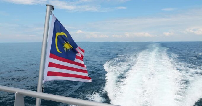 Malaysian national flag flutters on upper deck of ferry catamaran bound for Tioman Island. Vessel's powerful engines carve a wide, foamy white wake across the calm bluer waters
