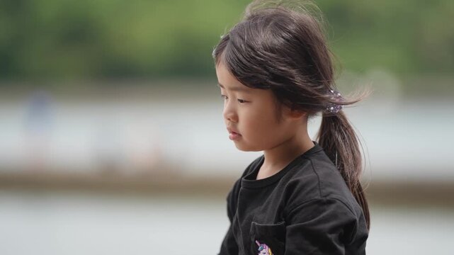 Child stands by calm water, wind lifts hair. Girl wears black top, focused on task ahead. Background blurred, greenery and water frame scene. Peaceful moment by natural setting, quiet and gentle