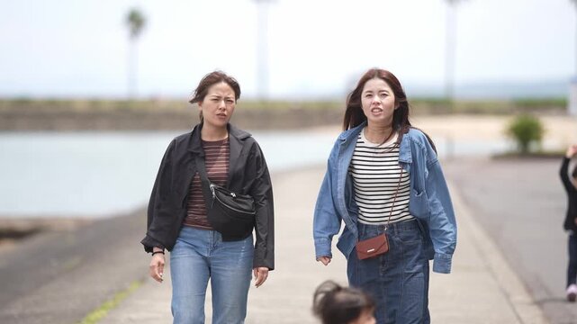 Two women walk along a coastal path. One wears a black jacket, the other a denim one. They carry crossbody bags and walk side by side. The scene feels relaxed, casual, and carefree