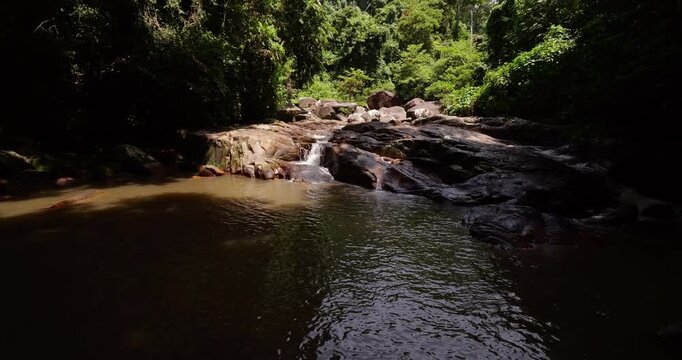 Shaded brown pond in foreground, with gentle stream cascading over rounded boulders, surrounded by dense tropical greenery. Camera tilts up to show Lubuk Teja Waterfall, Tioman Island local landmark