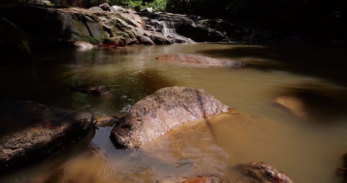 Sunlight illuminates pond at Lubuk Teja Waterfall, revealing its brown fresh water caused by suspended soil particles after rain. Camera slowly tilts up to reveal gentle stream