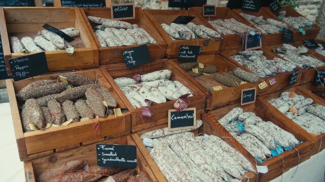 Medium High-Angle Shot of an Abundant Selection of Traditional French Saucisson Sec Displayed in Wooden Crates with Chalkboard Labels at an Outdoor Market in the South of France