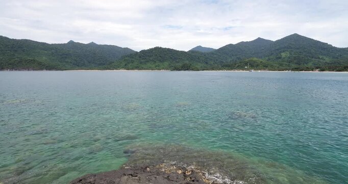 Panoramic view of Juara Beach on Tioman Island from sea rocks of small islet, with pristine turquoise waters stretching to shoreline, framed by verdant mountain slopes that surround picturesque bay