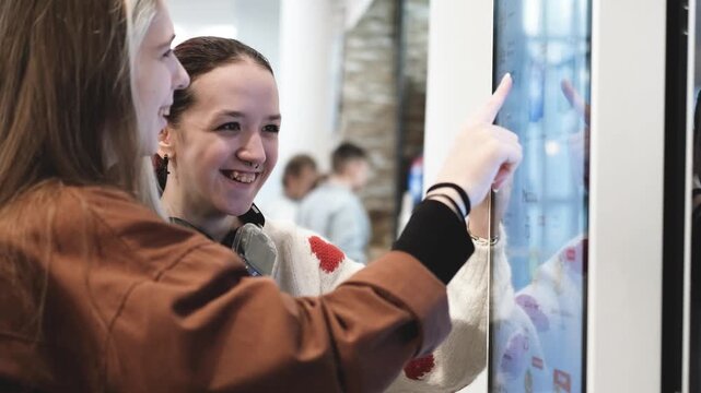 Two young women choose food from a menu on a self-service kiosk screen in a fast food restaurant.