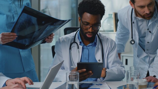 Professional multiethnic medical team of experienced doctors discussing x-ray using digital tablet laptop talking about diagnosis healthcare briefing three medicine workers talk brainstorm at hospital