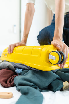 Close-up of Asian woman sitting on an overpacked yellow suitcase to close it. Concept of overpacking issues, travel preparation struggles and holiday planning. Vertical Shot