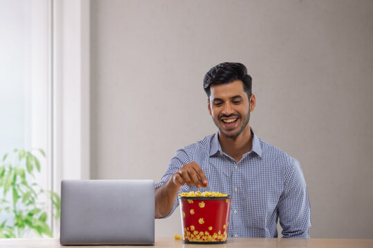 A happy corporate employee in formal clothing sitting with a laptop eating popcorn.