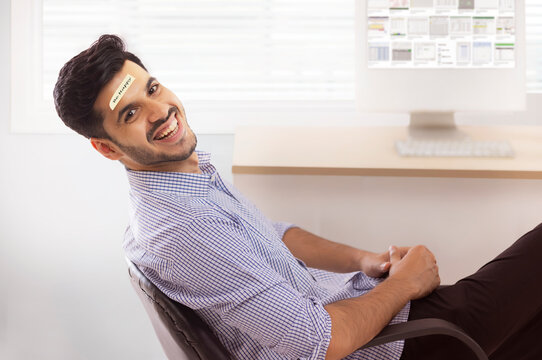 A corporate employee sitting in formal clothing smiling with stickynote on forehead.