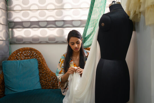 A young woman sitting and fixing her outfit on a mannequin with a  pin in mouth.