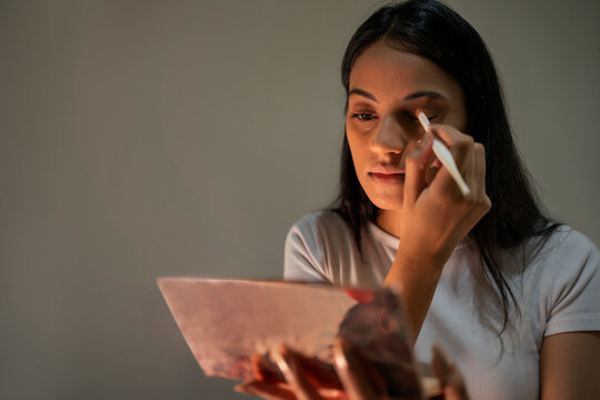 A young woman applying eye-shadow on her lids with a brush against plain background.