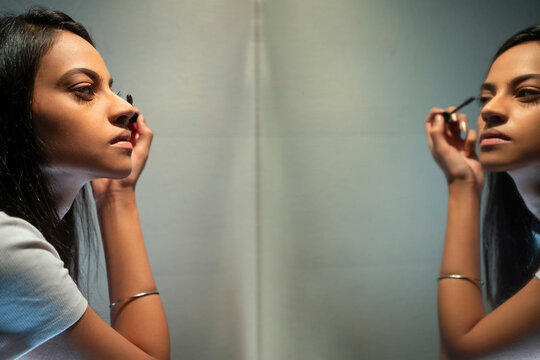 A young woman applying mascara with a wand sitting in front of mirror .