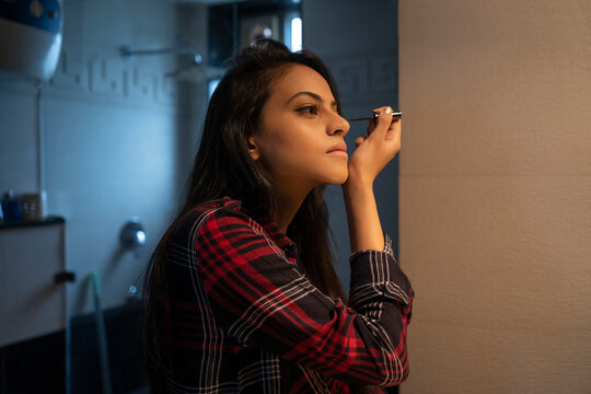 A young woman applying eyeliner on her lids with a wand while standing in the washroom.